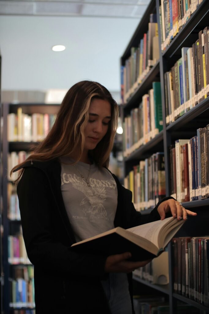 A young woman reading a book in a calm library setting, surrounded by shelves.